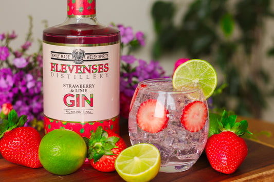 A bottle of Elevenses Strawberry and Lime Gin next to a refreshing summer cocktail, garnished with fresh strawberries and limes